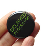 A round black pinback button that reads Delayed Processor in a lime green vintage digital text. Button is held in a hand.