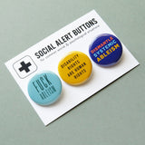 Three round pinback buttons lined up on a SOCIAL ALERT BUTTONS backing card. Reading left to right: A round aqua button reads FUCK ABLEISM, A yellow button that reads DISABILITY RIGHTS ARE HUMAN RIGHTS in black text, and a blue button that reads DISMANTLE SYSTEMIC ABLEISM in red, light blue & yellow.
