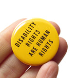 Round, golden yellow pinback button reads: Disability Rights are Human Rights in san serif capital letters. Button is held in a hand.