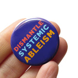 Round, dark blue pinback button that reads DISMANTLE SYSTEMIC ABLEISM in three lines in 3 different colors, red, light blue & yellow. Button is held in a hand.