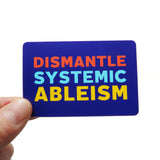 A purple blue rectangular sticker that reads DISMANTLE SYSTEMIC ABLEISM in three lines in neon red, light blue and yellow text. Held up by a hand.