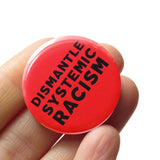Round pinback button that says DISMANTLE SYSTEMIC RACISM. Black text on a neon red background. Button is held in a hand.