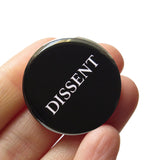 A round black pinback button that reads DISSENT in white serif text. Button is held in a hand.