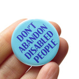 a round 1.25" pinback button with a pale aqua background that reads  DON'T ABANDON DISABLED PEOPLE in a blue-purple font. Button is held in a hand