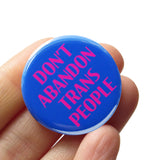 A round blue pinback button that reads DONT ABANDON TRANS PEOPLE in magenta pink. The button is held in a hand.