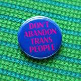 A round blue pinback button that reads DONT ABANDON TRANS PEOPLE in magenta pink. The button is on a green & blue patterned waffle knit shirt.