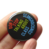 A round black pinback button with lime green. rust read and blue  text that reads SDONT STOP TALKING ABOUT CLASS CONSCIOUSNESS. The Button is held in a hand.