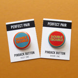 Two pinback buttons in inverse blue, orange and yellow colors that reads DOUBLE TROUBLE, each on a Perfect Pair backing card that perforates and tears down the middle to split the buttons, Keep One, Share One.
