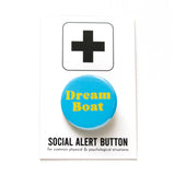 A round light blue pinback button that reads DREAMBOAT in two lines in yellow text. Button is on a white backing card that reads SOCIAL ALERT BUTTON in black text with a plus sign at the top.