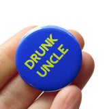 Round blue pinback button that reads DRUNK UNCLE in lime green text. The button is held in a hand.