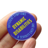 A round blue-purple 1.25" pinback button that's readsDYNAMIC DISABILITIES in neon yellow in the center, and DIFFERENT SYMTOMS & SEVERITY EACH DAY in smaller aqua text at the top & bottom. 