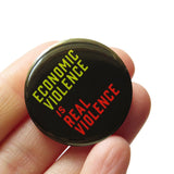 A round black 1.25" pinback button that reads Economic violence Is Real Violence. Badge is held in 