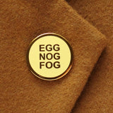 A round buttery pale yellow enamel pin with gold colored text that reads EGG NOG FOG in three lines, and a clean gold circular outline. Pinned to a warm camel colored wool coat lapel.