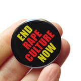 Round black pinback button that reads END RAPE CULTURE NOW in neon yellow and orange san serif text. The button is held in a hand.