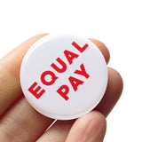 Round white pinback button that reads EQUAL PAY in red text. Button held in a hand.