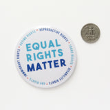 Big 3 inch round white pinback button with light blue & dark blue text that reads: TEQUAL RIGHTS MATTER. Smaller blue text runs in a circle around the permitter that says Reproductive Rights, Trans Rights, Disability Rights, Immigrant Rights, Voting Rights. Button is next to a quarter for size.