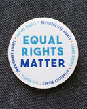 Big 3 inch round white pinback button with light blue & dark blue text that reads: TEQUAL RIGHTS MATTER. Smaller blue text runs in a circle around the permitter that says Reproductive Rights, Trans Rights, Disability Rights, Immigrant Rights, Voting Rights. Button is pinned to black denim
