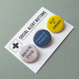 3 round multi-colored pinback buttons lined up on a SOCIAL ALERT BUTTONS backing card. Reading left to right: Brain Fog, Fatigue Fiasco, Out of Order