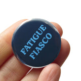 A deep muted blue pinback button that reads FATIGUE FIASCO in a lighter blue. Button is held in a hand.