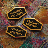 Three hexagon shaped hard enamel lapel pins that say FEMINIST CENTURY on a vintage colorful fabric