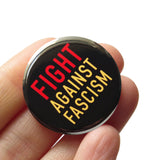 A round black pinback button that reads FIGHT AGAINST FASCISM in three lines in red and cream text. Button held in a hand.