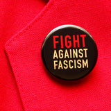 A round black pinback button that reads FIGHT AGAINST FASCISM in three lines in red and cream text. Button is pinned to a red blazer lapel.
