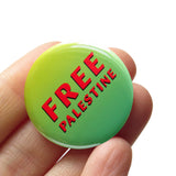 A round neon green ombre pinback button that reads FREE PALESTINE in 2 lines of red text. Button is held in a hand.