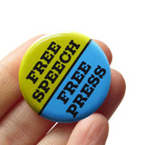 A round blue & lime green pinback button split down the middle. The green top half reads FREE SPEECH, the blue lower half reads FREE PRESS. Buton is held in a hand.