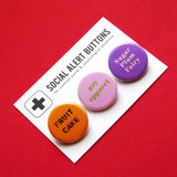 3 round pinback buttons lined up on a SOCIAL ALERT BUTTONS backing card. Reading left to right: Fruit Cake in Carmel colors, GAY APPAREL in lime green text on light pink button & SUGAR PLUM FAIRY in cream on lavender button. Buttons on card are on a red background.