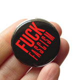 A round black pinback button that reads FUCK FASCISM in red text. Button held in a hand.
