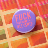 A round medium lavender pinback button with neon red text that reads FUCK Performative Allyship. Button is on an orange patterned surface