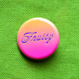 A peach to pink ombre pinback button that reads FRUITY in a purple scripty font with a slight neon yellow drop shadow.  Badge is pinned to a vivid green canvas bag.