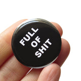 A round black button that reads FULL OF SHIT in white text. Badge is held in a hand.