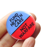 A round pinback button split between two colors, hospital scrubs blue on the top and neon Advil red-orange on the bottom. The top of the button reads: FUND HEALTHCARE, The bottom reads: NOT WARFARE. Badge is held in a hand.
