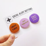 3 round pinback buttons lined up on a SOCIAL ALERT BUTTONS backing card. Reading left to right: Fruit Cake in Carmel colors, GAY APPAREL in lime green text on light pink button & SUGAR PLUM FAIRY in cream on lavender button. Buttons on card are on a mint green background.