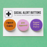 3 round pinback buttons lined up on a SOCIAL ALERT BUTTONS backing card. Reading left to right: Fruit Cake in Carmel colors, GAY APPAREL in lime green text on light pink button & SUGAR PLUM FAIRY in cream on lavender button. Buttons on card are on a mint green background.