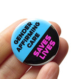 A round 1.25" pinback button with a blue and black split color design, reading "GENDER AFFIRMING CARE"in black text on the blue portion, and "SAVES LIVES" is in hot pink on the black background. Button is held in a hand.