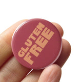 Round rosy strawberry muffin colored 1.25" pinback that reads GLUTEN FREE in golden text. Badge is held in a hand.