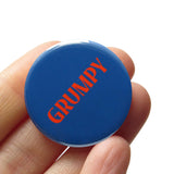 Dark navy blue round pinback button that reads GRUMPY in a red-orange font. Button is held in a hand.
