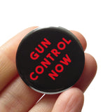 Round black pinback button that reads GUN CONTROL NOW in red text on three lines. Badge is held in a hand.