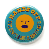 A round teal pinback button that reads HANDS OFF Civil Rights & Social Safety Nets yellow-orange text,  with an angry shouting face in the center of the button. 