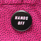 A round black pinback button that reads HANDS off in white text with a pink drop shadow. Pinned to an 80's design pink shirt with tiny triangles on it.