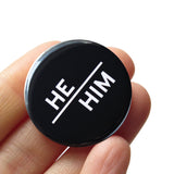 Round pinback button with black background which reads HE/HIM on an angle in white text, in a san serif font. Badge is held in a hand.