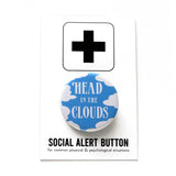A round medium blue 1.25" pinback button with white fluffy clouds along the edges and the words " HEAD IN THE CLOUDS" in a white art deco font on the face of the button. The button is on a white card with a black plus sign and the text 'SOCIAL ALERT BUTTON for common physical & psychological situations'.