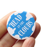 A round medium blue 1.25" pinback button with white fluffy clouds along the edges and the words " HEAD IN THE CLOUDS" in a white art deco font on the face of the button. The button is held in a hand.