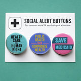 3 round multi-colored pinback buttons lined up on a SOCIAL ALERT BUTTONS backing card. Reading left to right: Healthcare Is a Human Right, Public Health Over Corporate Wealth, Save Medicaid