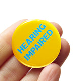 Round, yellow 1.25" pinback button reads: HEARING IMPAIRED in light blue text. Button is held in a hand.