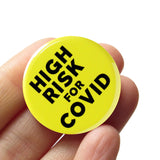 Round neon yellow 1.25" pinback button that reads HIGH RISK FOR COVID in chunky black text.  Badge is held in a hand.