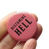 Round rosy mauve 1.25" pinback with splatter dots button reads HISTAMINE HELL in black text. Badge is held in a hand.