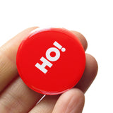 a round red pinback button that reads HO! in white text. Button is held in a hand.